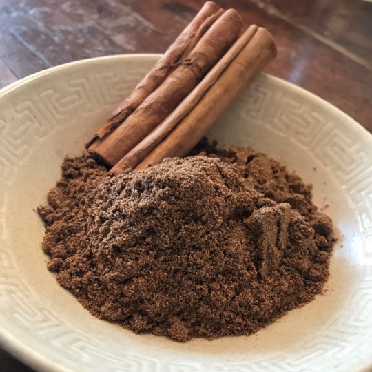 A little creamy white pale brown bowl holds a mound of brown spices on the top of the little bowl sits two cinnamon sticks